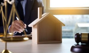 Wooden model home atop table with businessman in background with gavel and scale on table