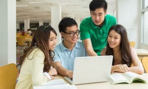 Group of 4 students grouped in library around laptop and textbooks