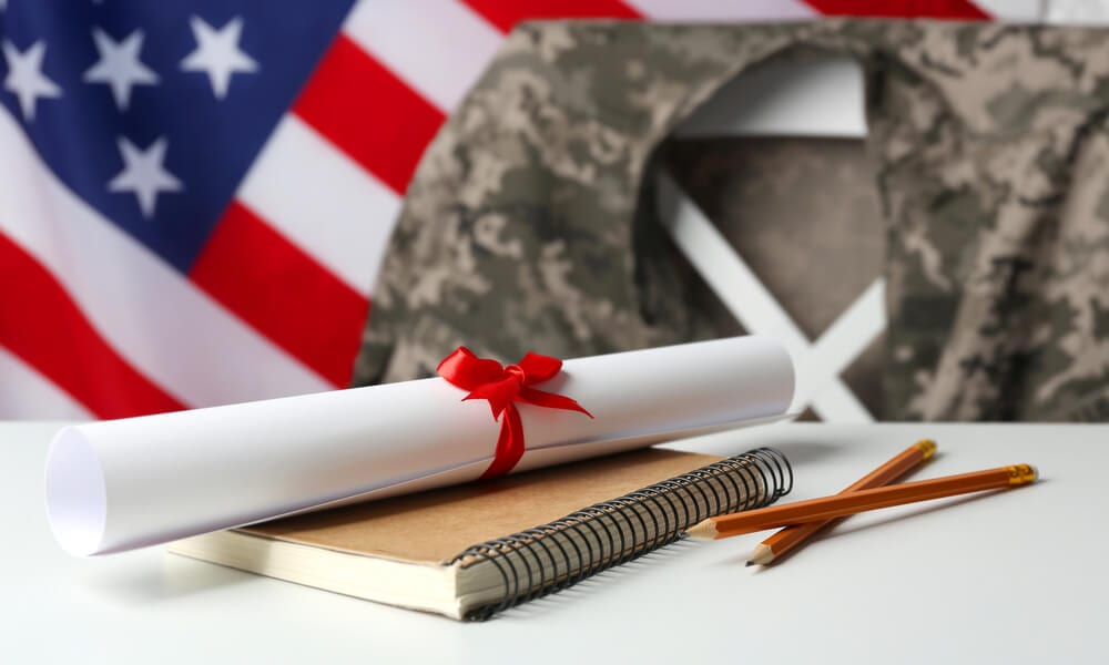 Notebook, diploma and pencils on white table with military jacket hanging on chair and American flag in background