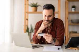 Man sitting at table in home office, holding and looking at credit card considering debt relief options.