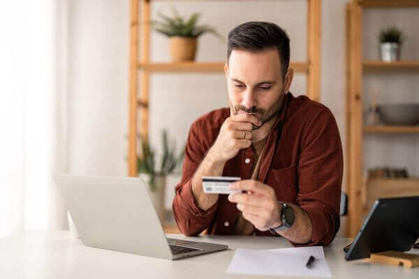 Man sitting at table in home office, holding and looking at credit card considering debt relief options.