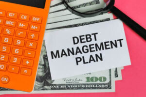 Phrase DEBT MANAGEMENT PLAN written on white card with magnifying glass, fake money and calculator.